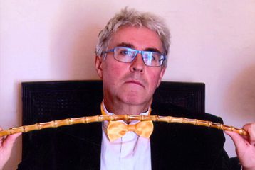 An image of Peter Freeman wearing a bow tie and a suit, and holding a stick between his hands.
