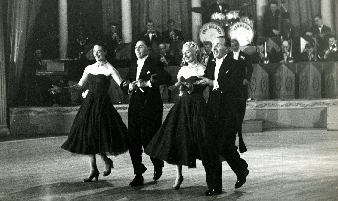 Two men and two women dancing, with a band behind them. The women are wearing strapless black dresses and the men are wearing suits.