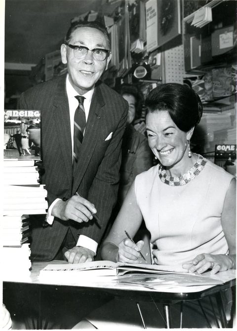 A photo of Peggy writing, and her husband Frank standing behind her.