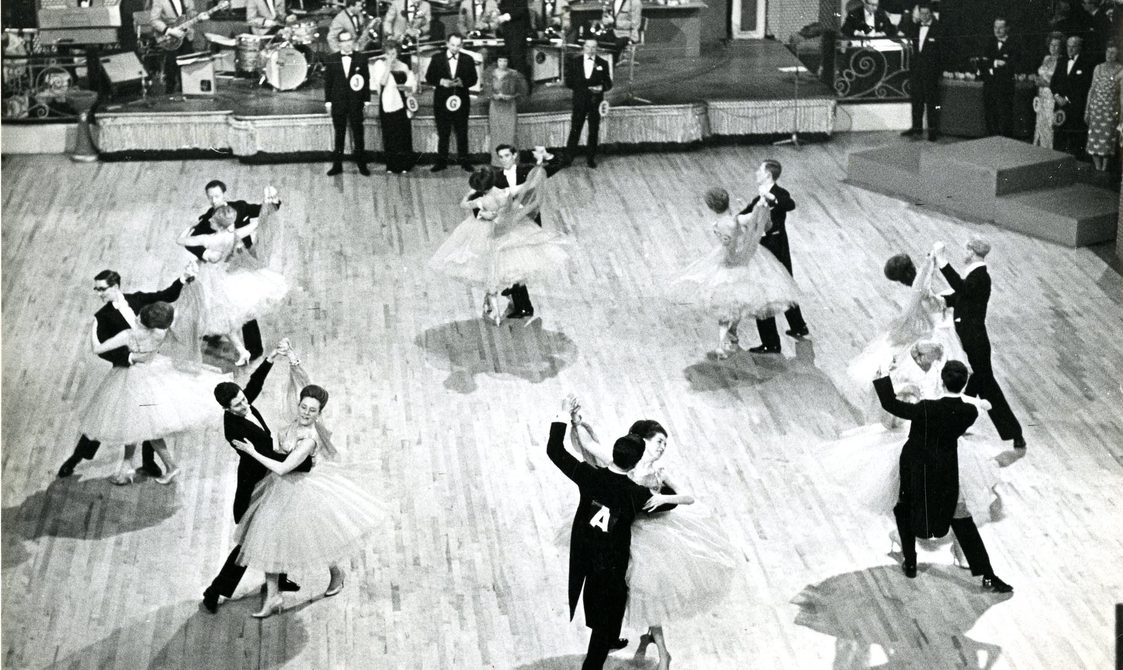 Eight couple on a dance floor, twirling round. They are in a circle as they move. A band is playing behind them.