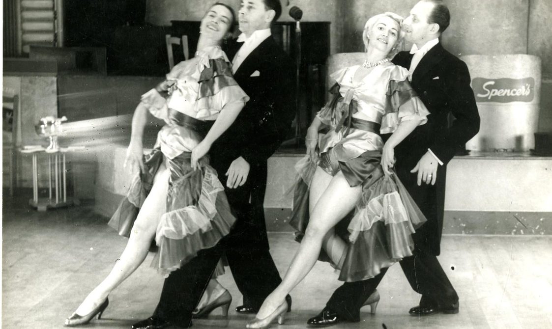 Two male dancers and two female dancers in front of a stage. The female dancers are standing in front of the male dancers, with their right leg placed in front of their left, and they're wearing flamingo style dresses.