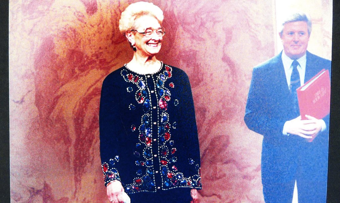 A photo of Peggy Spencer wearing a blue shirt and glasses and smiling. A man is standing behind her in a suit, and holding a red book.