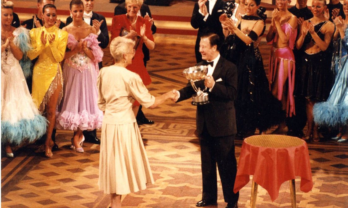 Peggy Spence being given a trophy in 1990 at Blackpool, surrounded by dancers clapping.