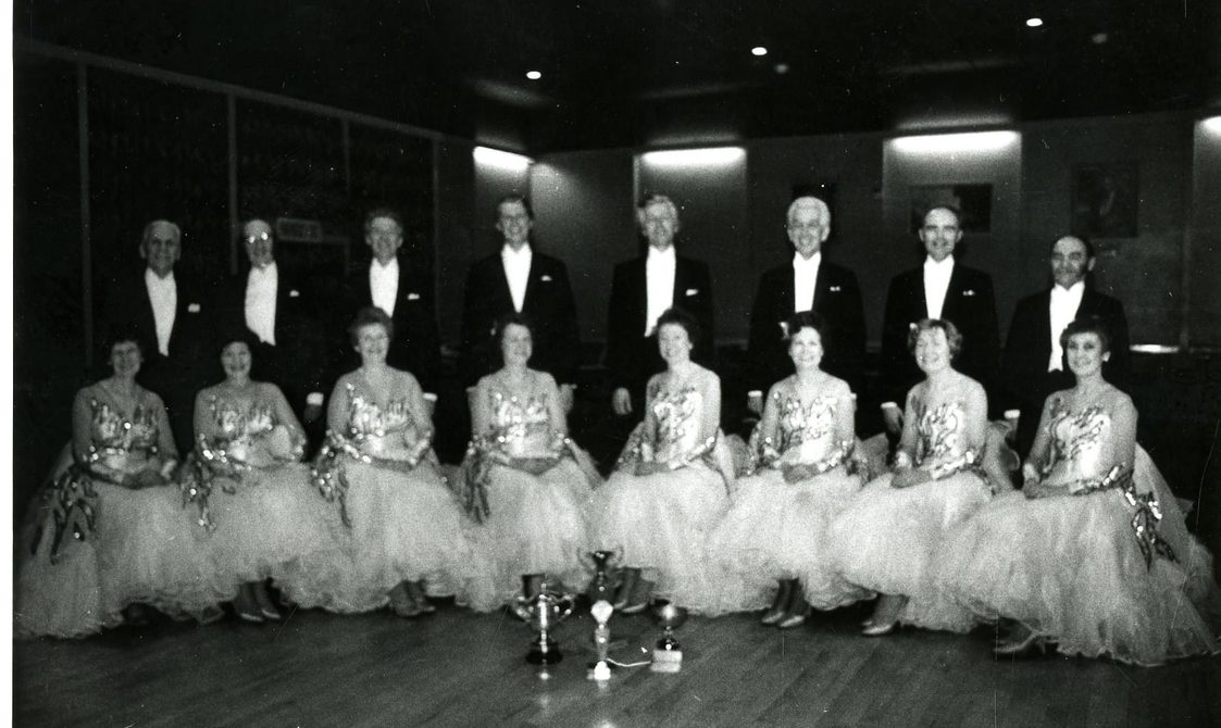 Nine dancing pairs kneeling on a ballroom floor, with the trophy in front of them.
