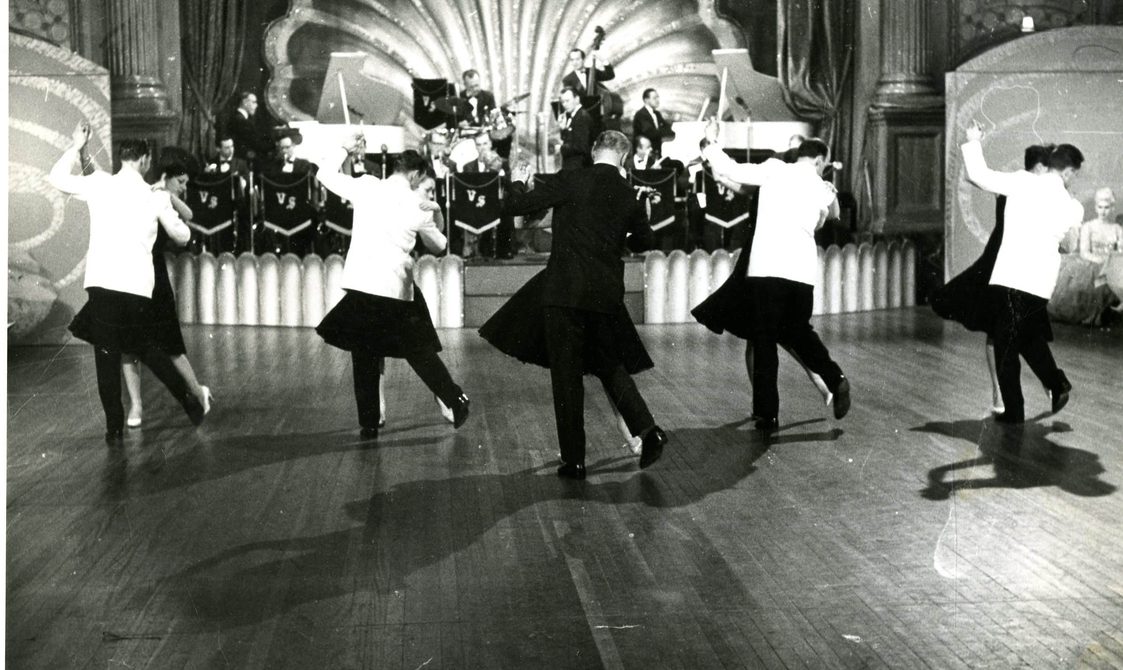 A photo of the Penge Latin team performing on stage in 1963. A band is behind them and five couples are on stage,