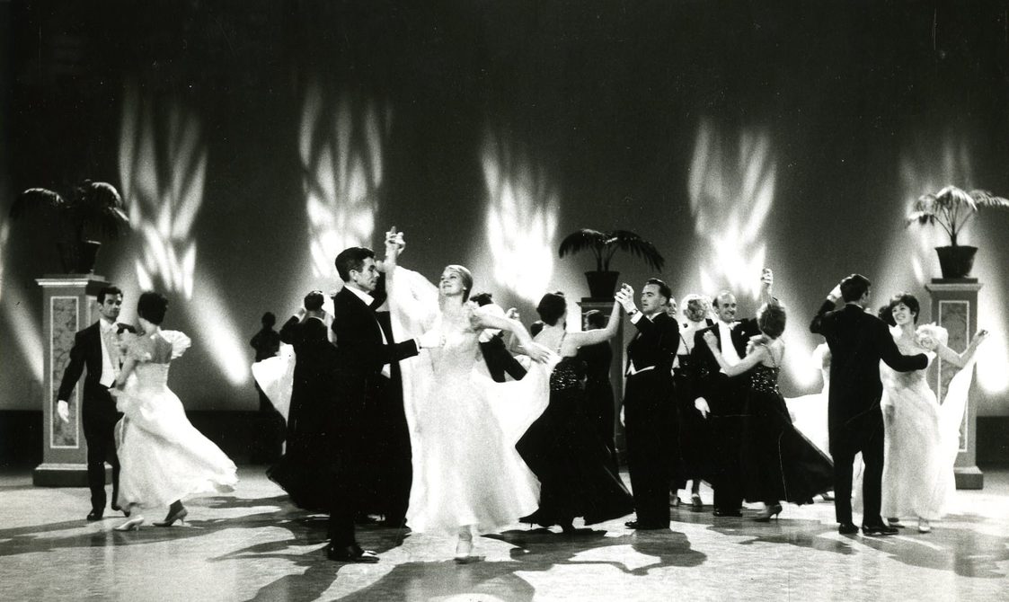 Dancers performing on stage as part of Dancing Years. The men are in suits and the woman in white long dresses, and they performing as couples.