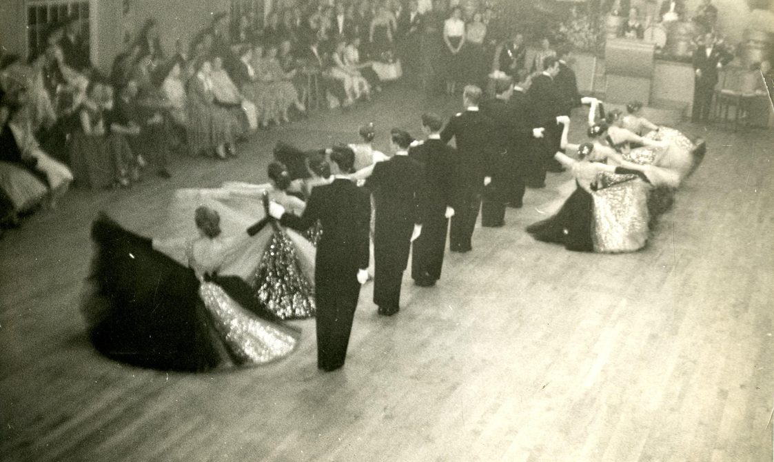 A black and white photo of the original Penge team, led by Peggy Spencer, performing. The woman are curtsying on the dance floor with the men standing next to them. An audience is seated around them.