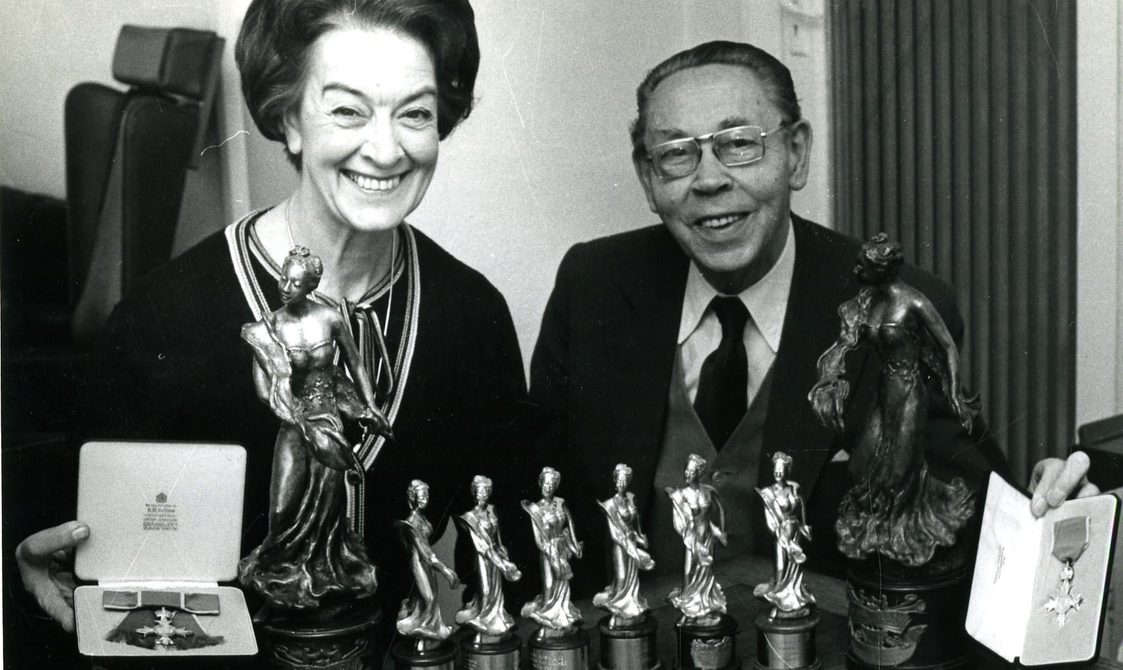 Peggy Spencer and her husband Frank sitting down with various trophies in front of them.