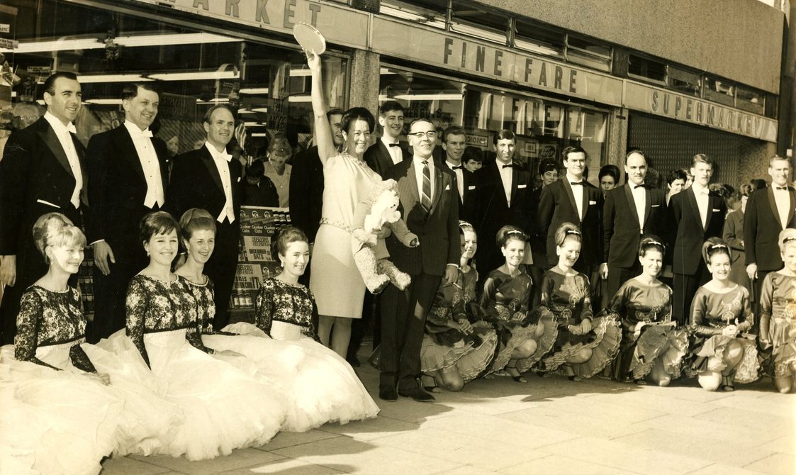 A sepia photo of dancers. The girls are kneeling on the floor, with the men standing behind them. In the middle is Peggy Spencer waving.
