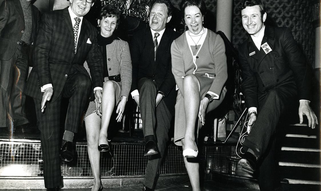 A picture of four people dancing on stage, with their left legs up in the air, and clapping underneath their legs. They are smiling and looking at the camera.