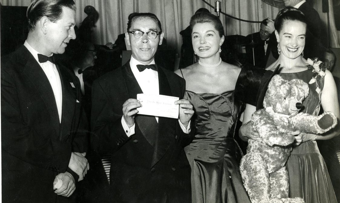 Peggy Spencer with Esther Williams. Peggy is holding a teddy bear and smiling at the audience, as she stands on stage.