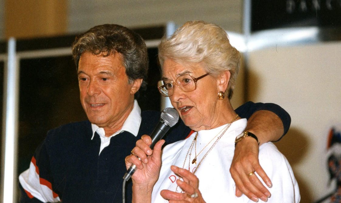 A picture of Peggy Spencer speaking into a microphone, with Lionel Blair next to her, who has his arm around her.