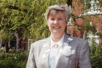 A photograph of Sue King, smiling. She has short hair and is wearing a grey plaid suit.