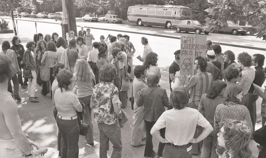 A picture from the second Pride march in 1973. A group of people are standing around Embankment before the march starts. One person is holding a sign that says: "Gay Liberation Front".