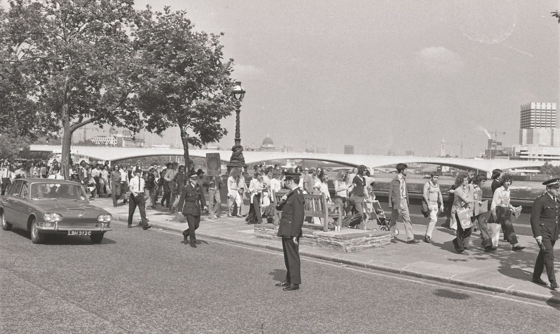 A picture from the second Pride march in 1973. A group of people are marching down from Embankment to Hyde Park. Police officers are around the marchers.