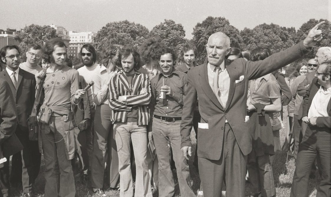 A picture from the second Pride march in 1973. A group of protestors are in Hyde Park, with a man at the front pointing to his left as he addresses the protestors.