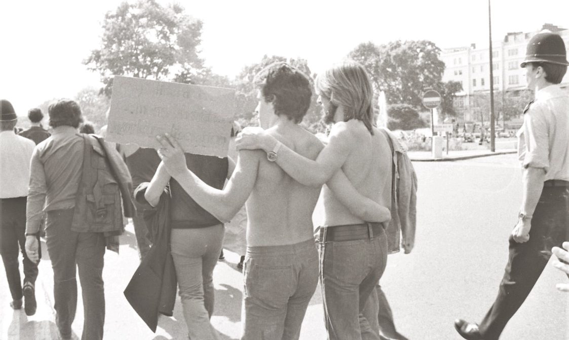 A picture from the second Pride march in 1973. Marchers are walking with their arms around each other, and police standing nearby.