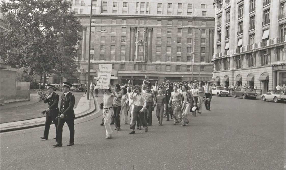 A picture from the second Pride march in 1973. Marchers are walking down the road, with police in front. The people at the front are holding a sign that says: "Young homosexuals have the right to love".