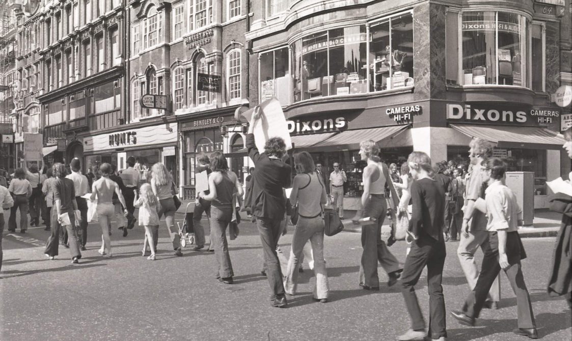 A picture from the second Pride march in 1973. Marchers are walking down the road in Central London, going past the shop Dixons.