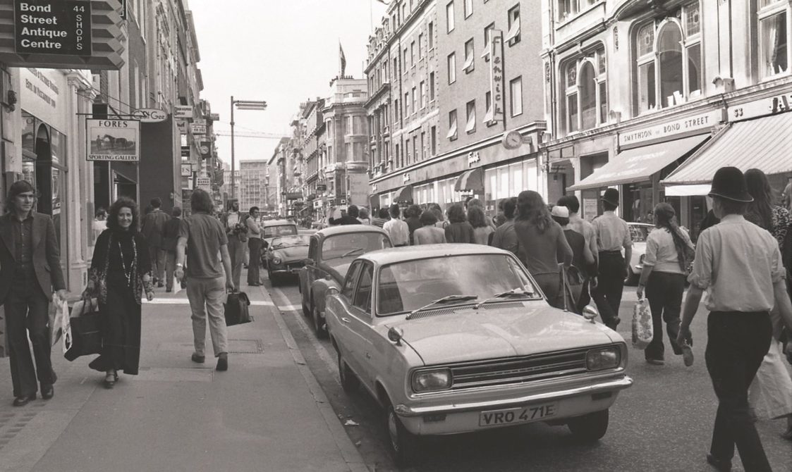 A picture from the second Pride march in 1973. Marchers are walking down the road in Central London, where cars are parked.