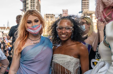 Two people are looking at the camera in the middle of a march. The on eon the left has long blonde hair, is wearing a dress of the trans pride colours and a trans pride mask. The other is wearing a silver sequin tops and glasses.