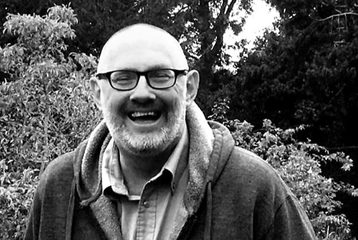 A black and white photo of a smiling person with glasses and beard wearing a hoodie, standing outdoors in a garden.