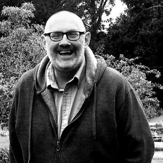 A black and white photo of a smiling person with glasses and beard wearing a hoodie, standing outdoors in a garden.