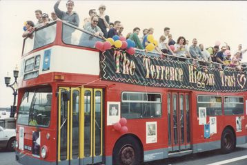 A red open top bus, with lots of people on top. There are balloons, and a bit banner reading "The Terrence Higgins Trust"