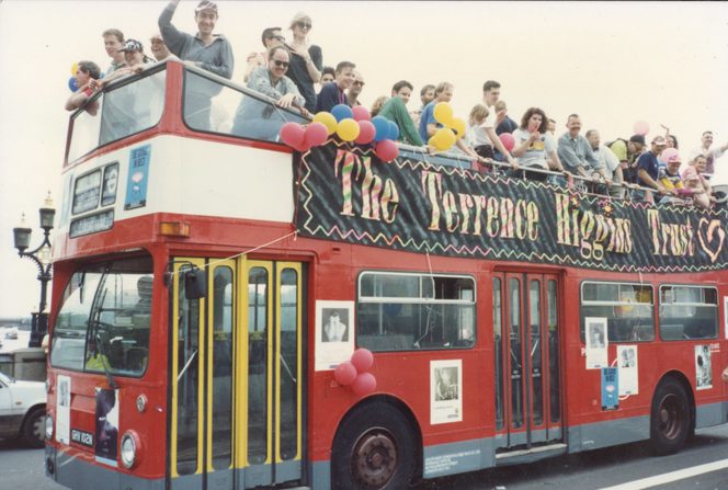 A red open top bus, with lots of people on top. There are balloons, and a bit banner reading "The Terrence Higgins Trust"
