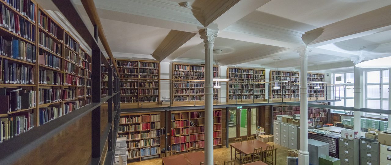 Wide view of Bishopsgate researchers area. Shelves full of books with desks and chairs in the middle.