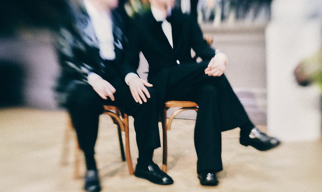 A couple in tuxedos sit and hold hands.