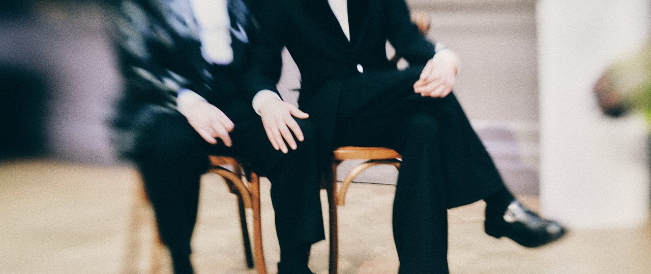 A couple in tuxedos sit and hold hands.