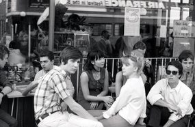 A black and white photograph of a group of young people sat at tables outside a cafe.