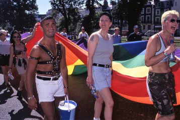 A group of people walking in the 1998 Brighton Pride parade, carrying a section of a giant rainbow flag that runs the length of the street.