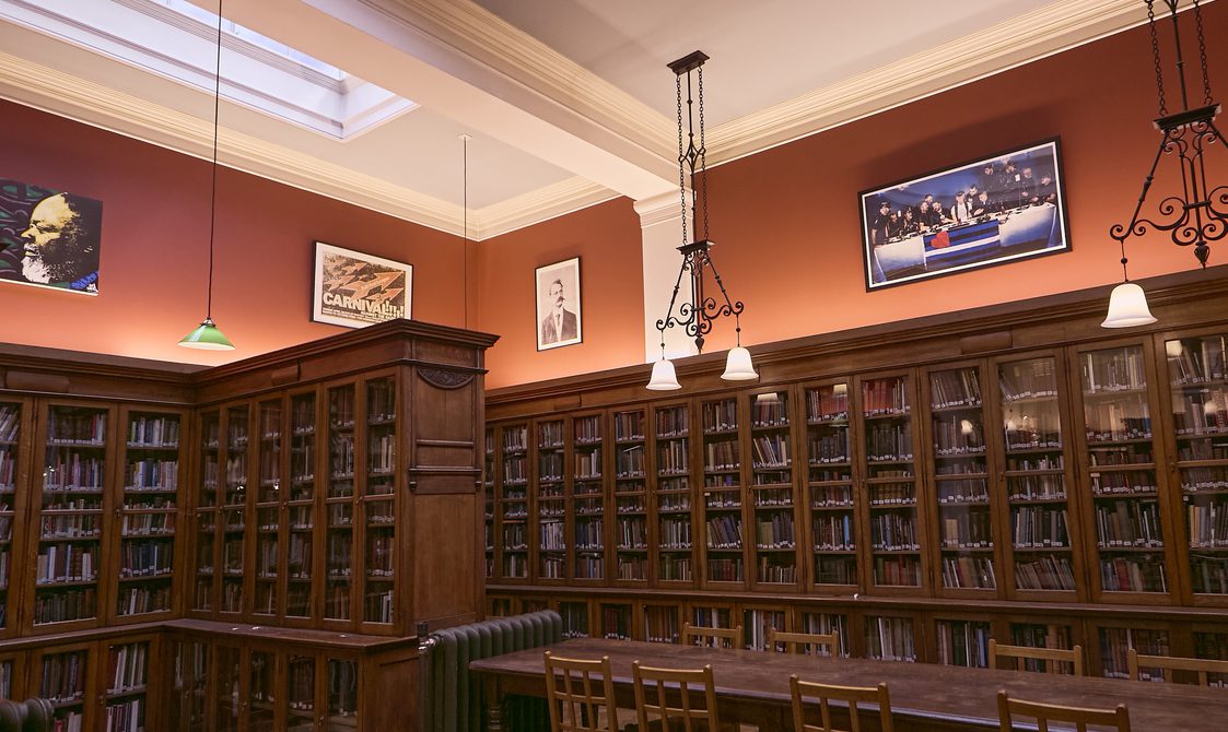 A landscape photo of Bishopsgate Institute Library. The floor is laminate wood flooring, with books in cabinets around the edges of the room.
