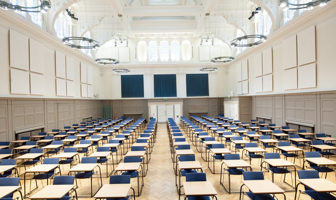 A photograph of Bishopsgate Institute's Great Hall in London. Single tables are set up in rows with blue chairs.