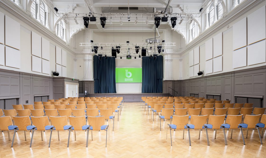 A photograph of Bishopsgate Institute's Great Hall in central London. The chairs are laid out in rows facing the stage, which has a digital screen with Bishopsgate Institute's logo on.