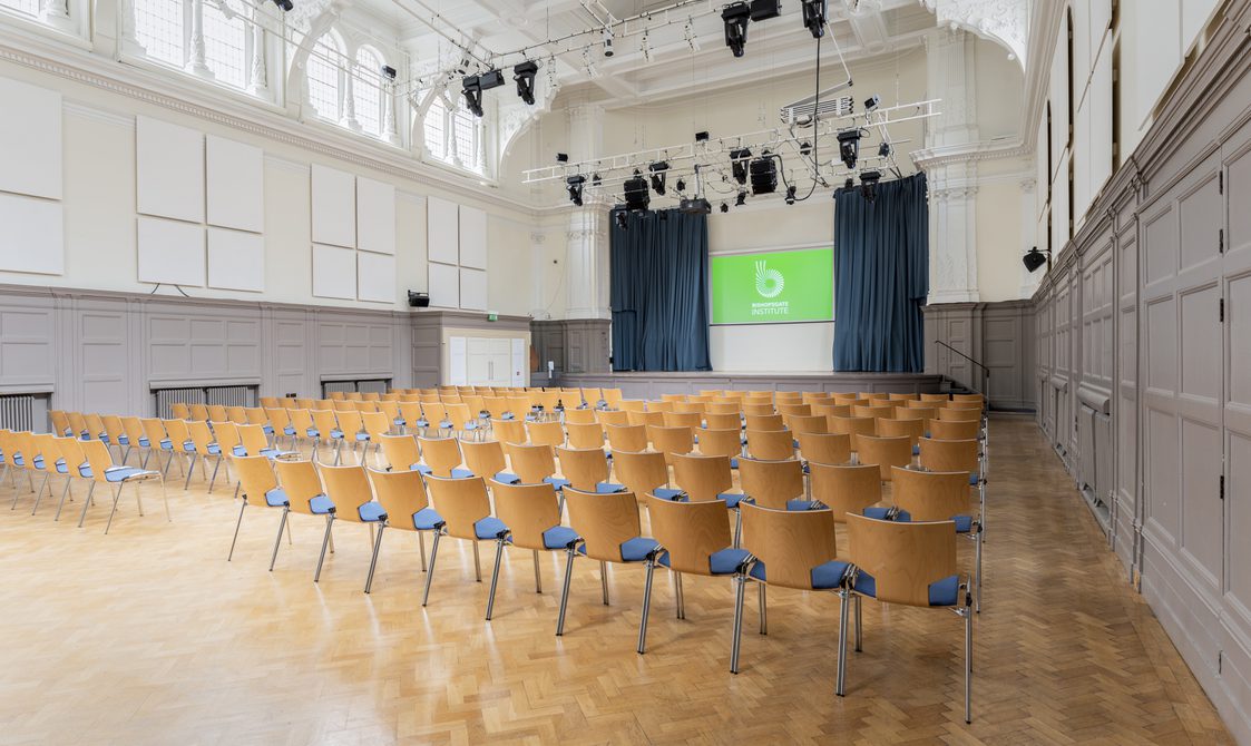 A photograph of Bishopsgate Institute's Great Hall in central London. Chairs are put out in rows facing the stage, which has a digital screen with a green background and Bishopsgate Institute's logo on it.