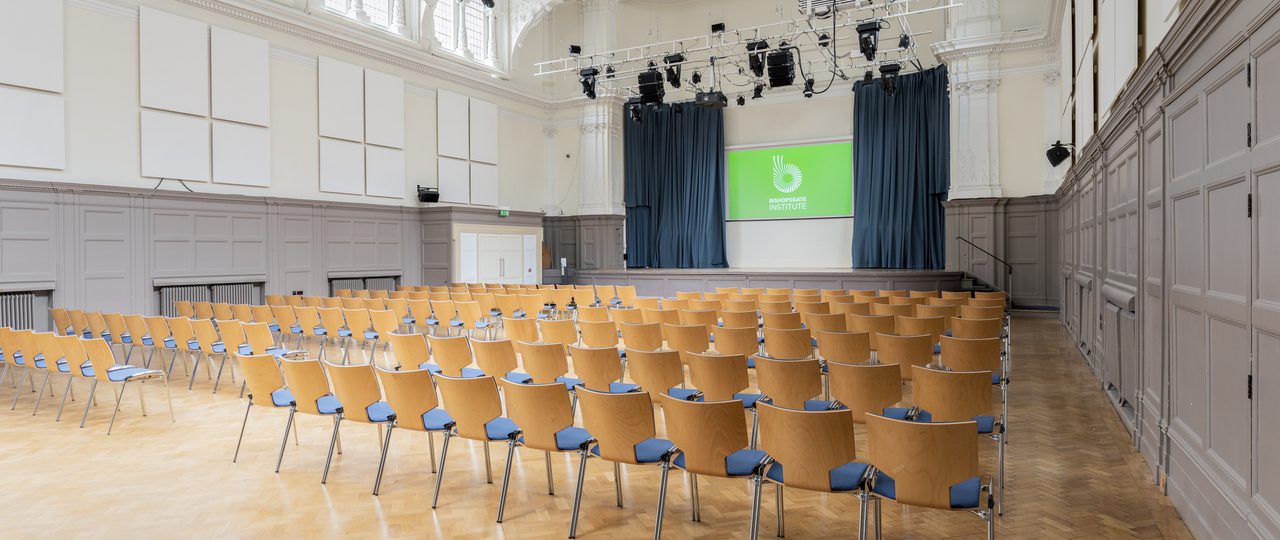 A photograph of Bishopsgate Institute's Great Hall in central London. Chairs are put out in rows facing the stage, which has a digital screen with a green background and Bishopsgate Institute's logo on it.