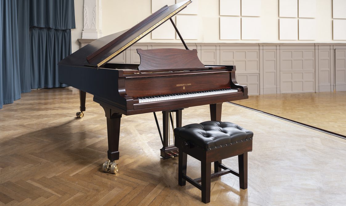 A photograph of a piano on stage at Bishopsgate Institute in London. The piano is a Model D Steinway that Myra Hess performed on.
