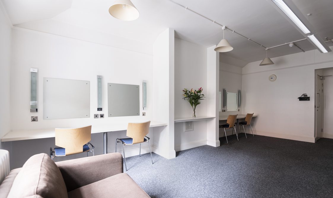 A photograph of Bishopsgate Institute's Green Room in central London. The floor is carpeted and there are chairs facing mirrors, with flowers on the table. There is also a sofa in the room.