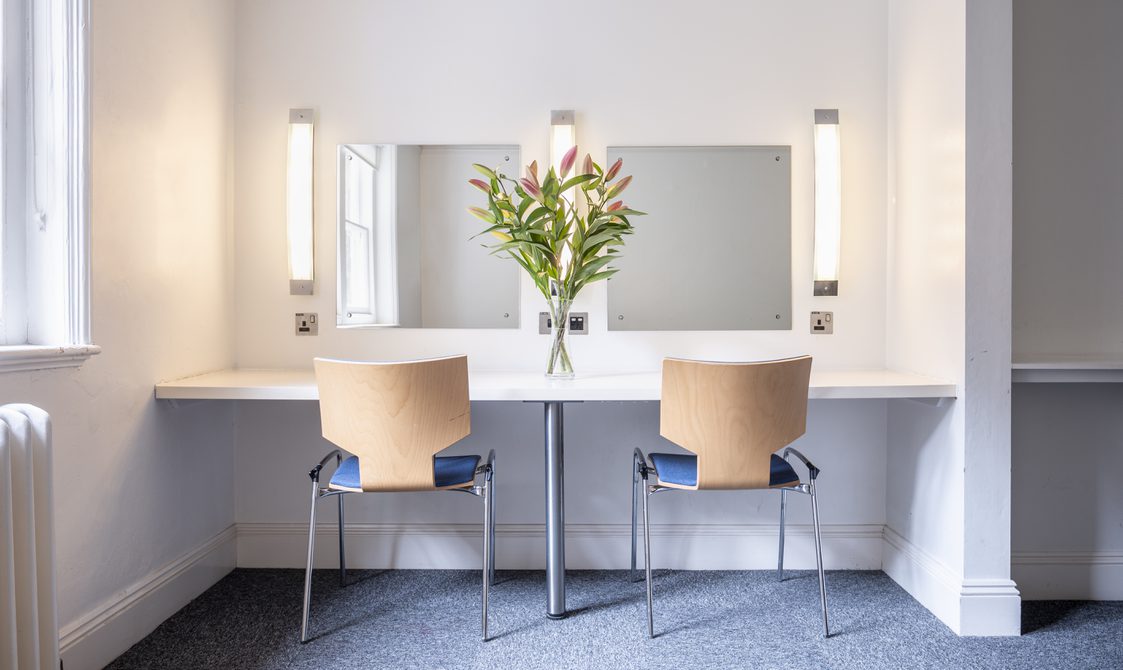 A photograph of Bishopsgate Institute's Green Room in central London. Two chairs are by a white table, which has a mirror in front of it with flowers on the table. It has been done in a dressing room style.