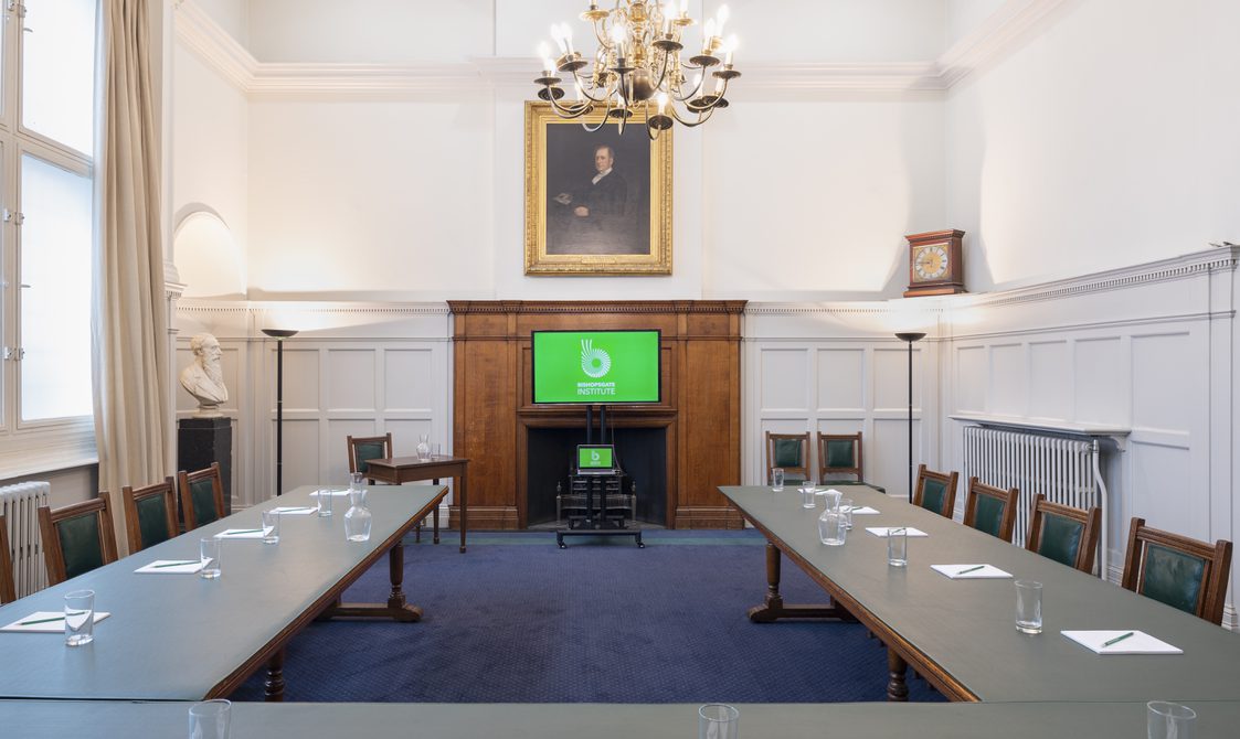 A photograph of Bishopsgate Institute's Boardroom in central London. The tables are set out in a square with chairs around them. There is a fireplace with a screen in front of it. There is a portrait hanging with a chandelier from the ceiling.