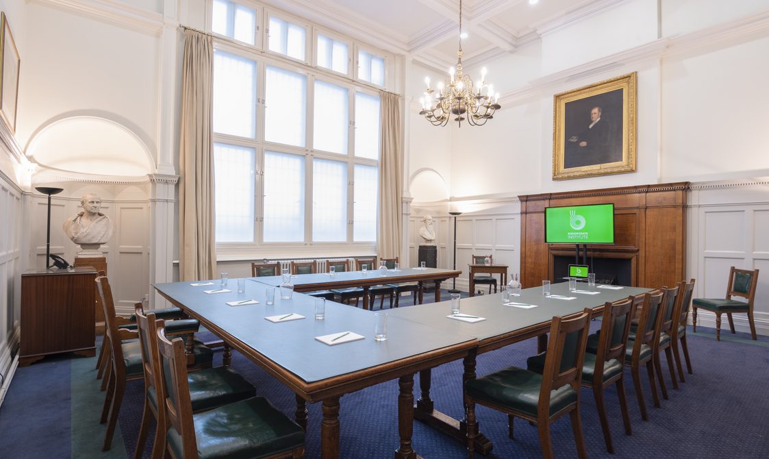 Tables are in a U-shape with chairs around them. There is a fireplace at the front of the room with a digital screen in front of it. Above it is a portrait and the walls are white with a chandelier hanging from the ceiling.