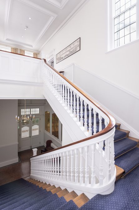 A photograph of the staircase leading up the Boardroom Suite at Bishopsgate Institute in central London. The staircase is white with a wooden handle and the stairs are carpeted blue.