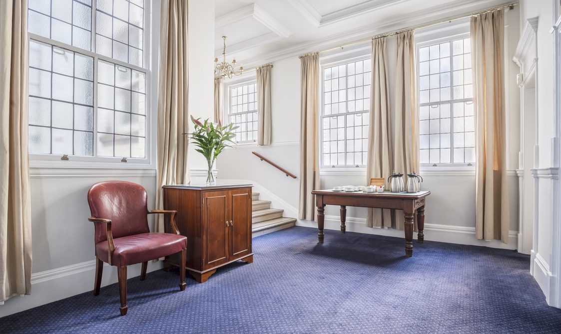 A photograph of the landing boardroom suite at Bishopsgate Institute in central London. The floor is carpeted with a wooden desk, chest of drawers, and a chair. There are then stairs leading up to the boardroom.