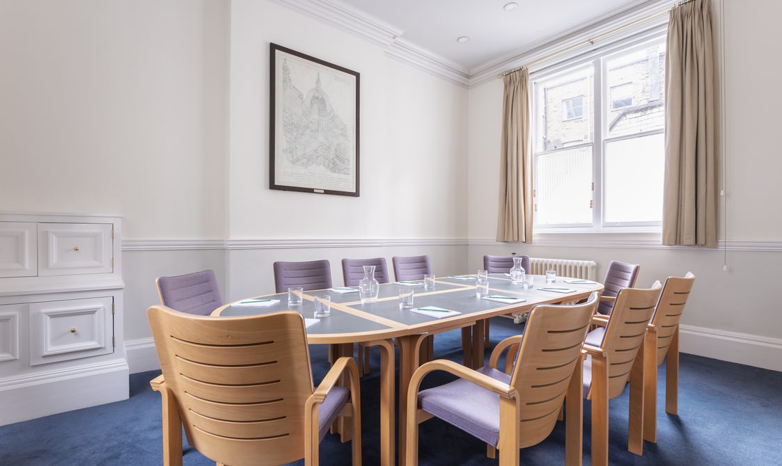 A photograph of the Townsend Room at Bishopsgate Institute in central London. The table is glass with ten chairs around it that have wooden backs with purple cushions. The walls are white with beige curtains by the window. There is an illustration on the wall facing the table.