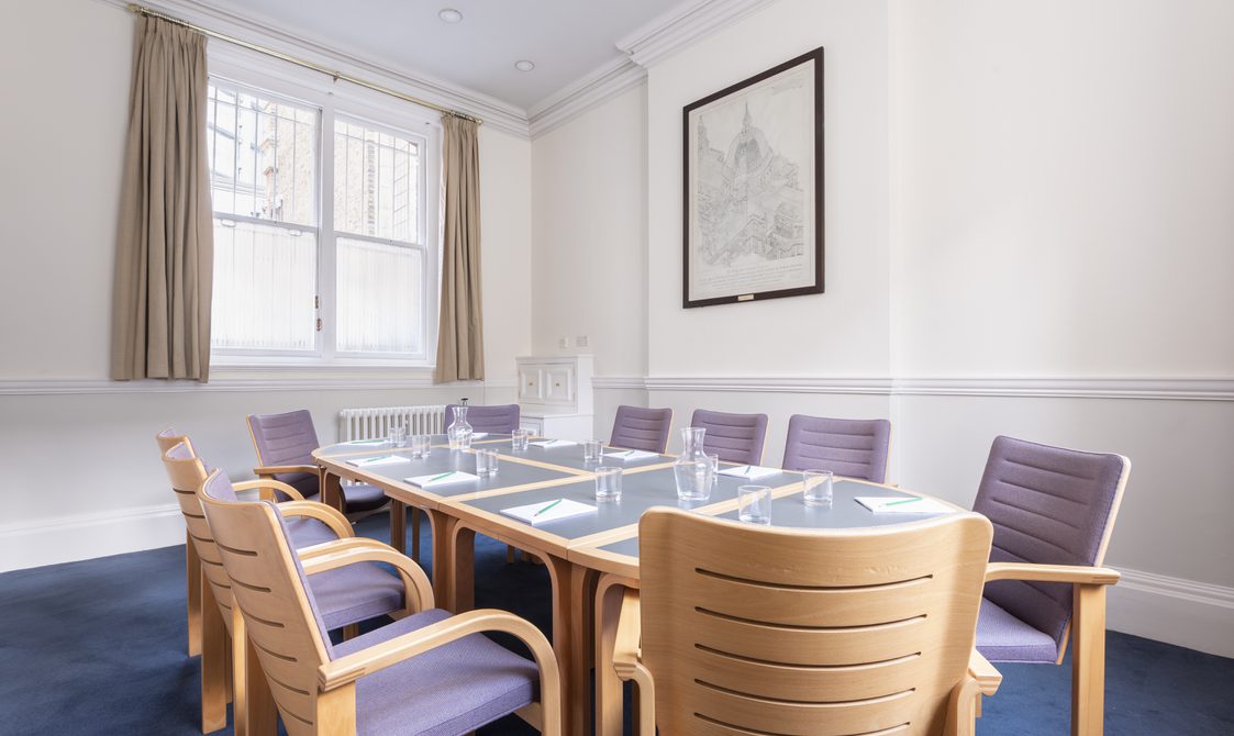 A photograph of Bishopsgate Institute's Townsend Room in central London. The table is set up in a boardroom style with chairs around it. The chairs are wooden with purple covers. The walls are white with a picture hanging on the wall and a window behind the table.