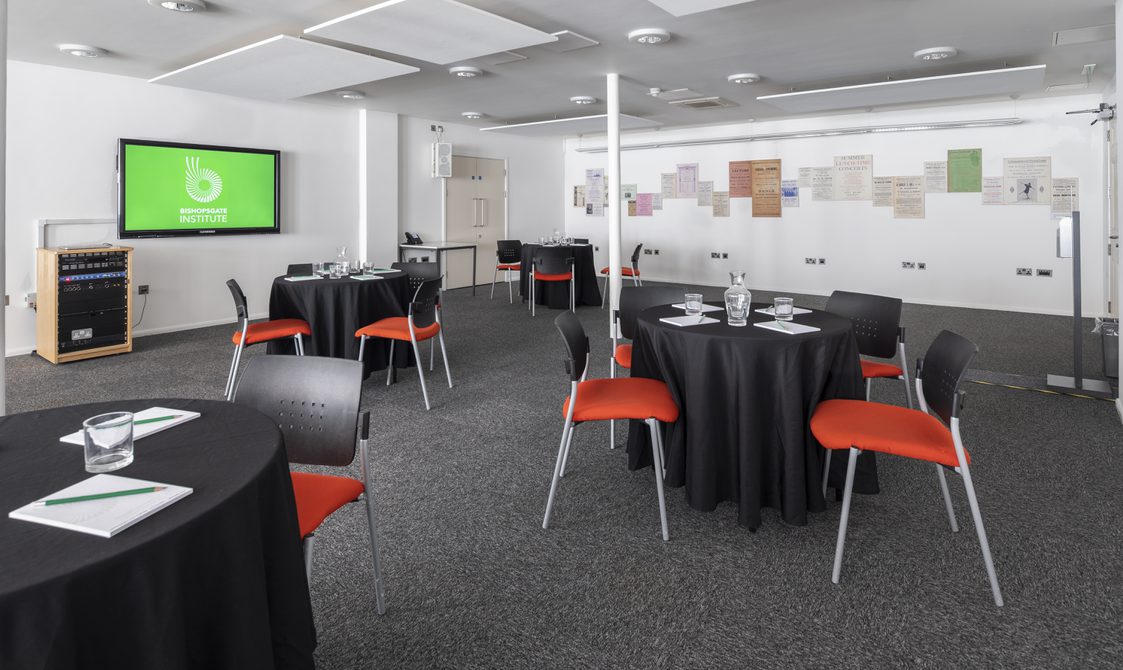 A photograph of Bishopsgate Institute's Courtyard Room in central London. The tables are set up in a cabaret style with chairs around them. A screen is mounted on the wall.