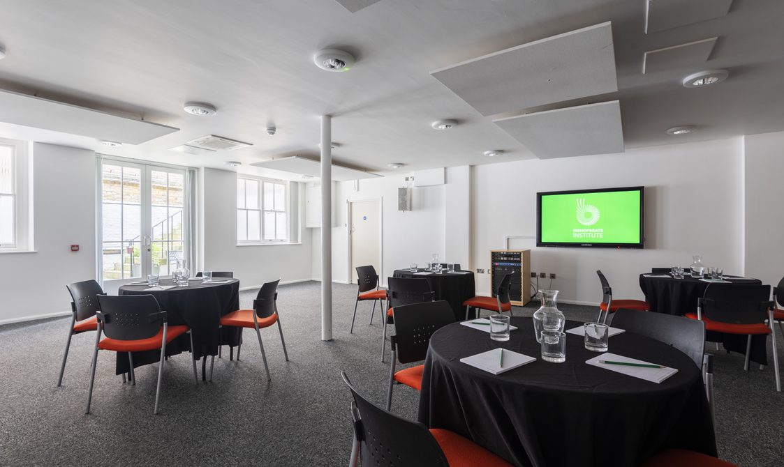 Four cabaret style tables are in the Courtyard room with a digital screen at the front of the room. The walls are white and there's a white beam in the centre of the room.
